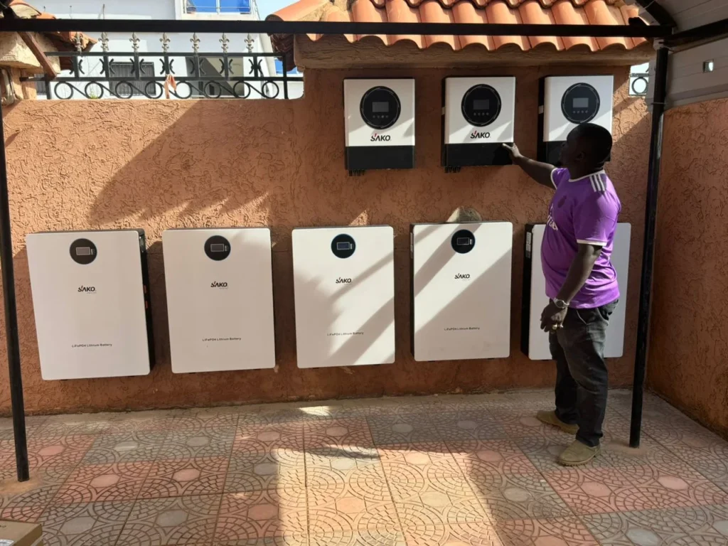 Installation of Sako LiFePO4 lithium batteries and solar inverters on a textured wall under a tiled roof, highlighting a display of renewable energy products.