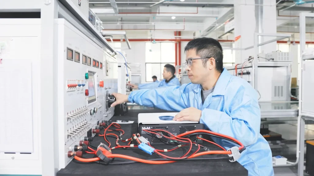 Engineer in blue uniform operates energy storage test system with cables on a desk, inside a manufacturing facility.