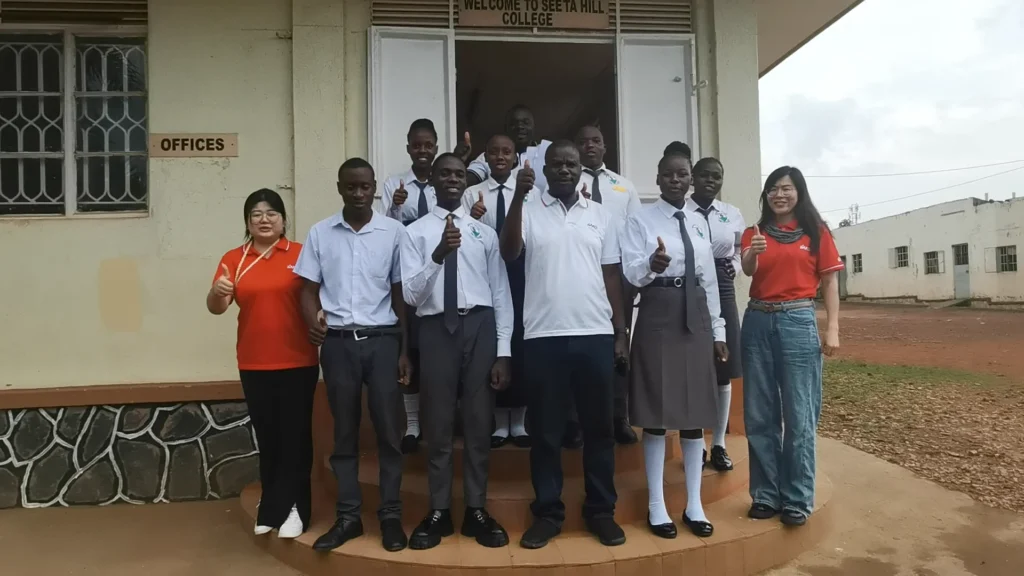 Group of people standing on steps at Seeta Hill College entrance, many giving thumbs up, wearing uniforms and casual clothes.
