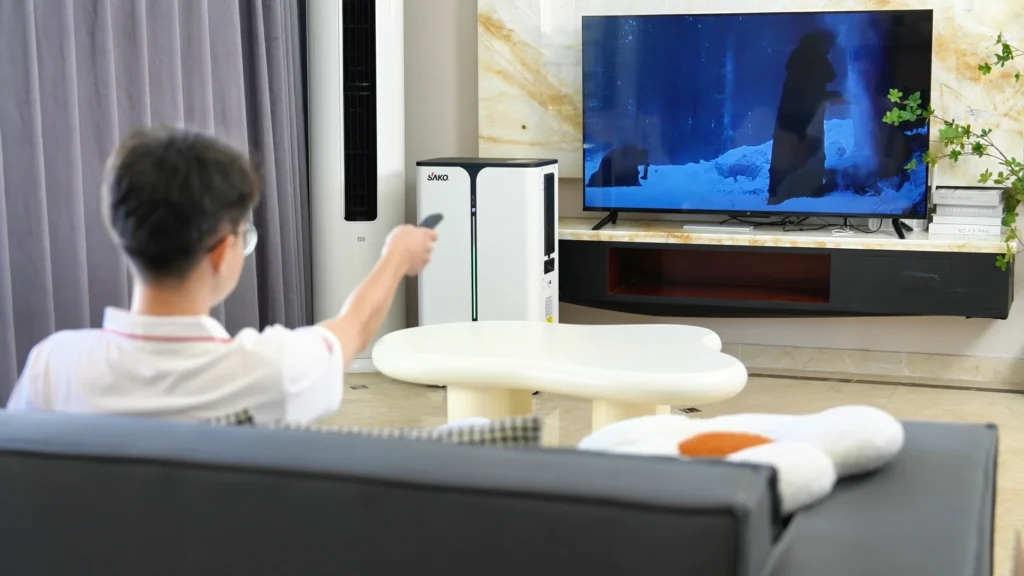 A modern living room featuring a SAKO energy storage system near a TV, with a person watching TV via a remote control and decorative elements around.