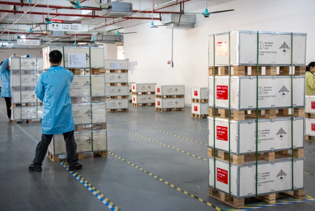 Workers in blue coats organizing stacks of packaged lithium batteries on pallets inside a factory setting with marked floors.