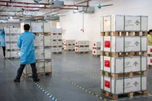 Workers in blue coats organizing stacks of packaged lithium batteries on pallets inside a factory setting with marked floors.
