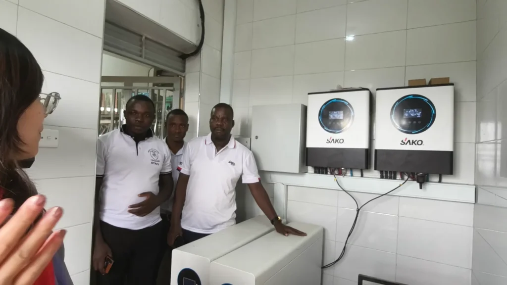Two SAKO solar inverters mounted on a wall, beside energy storage units, in a company setting with staff observing the equipment.