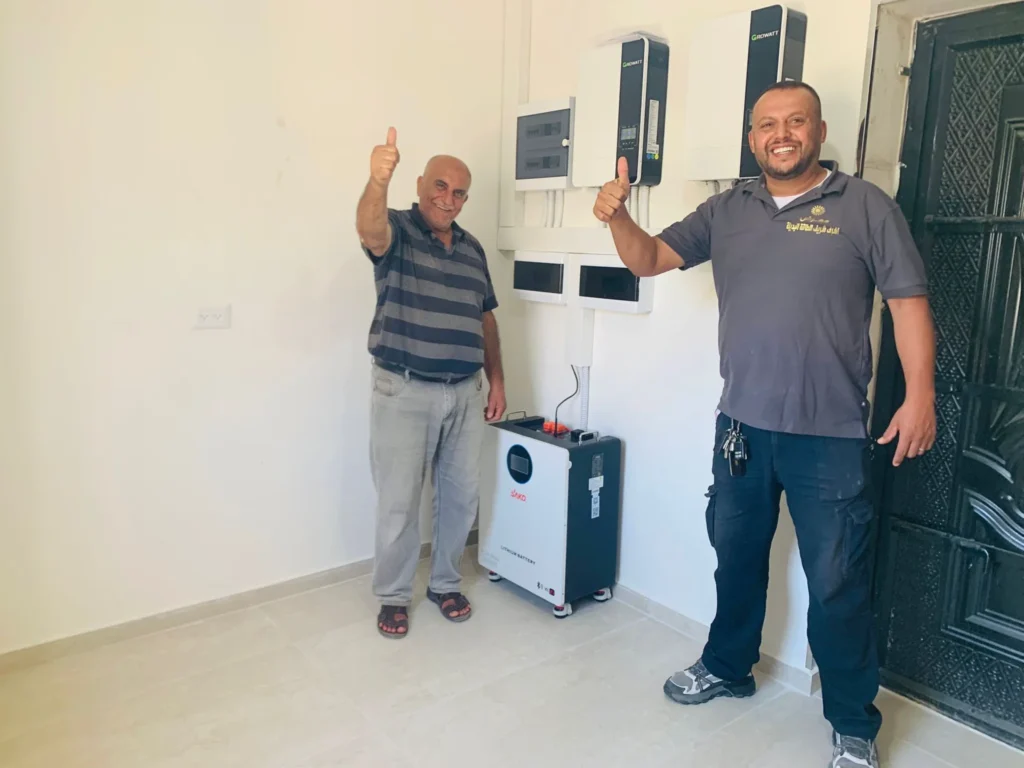Two men giving thumbs up in front of installed solar inverters and energy storage system inside a room.