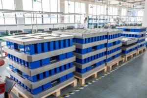 Stacks of blue battery packs on pallets in a factory assembly area with two workers in the background.