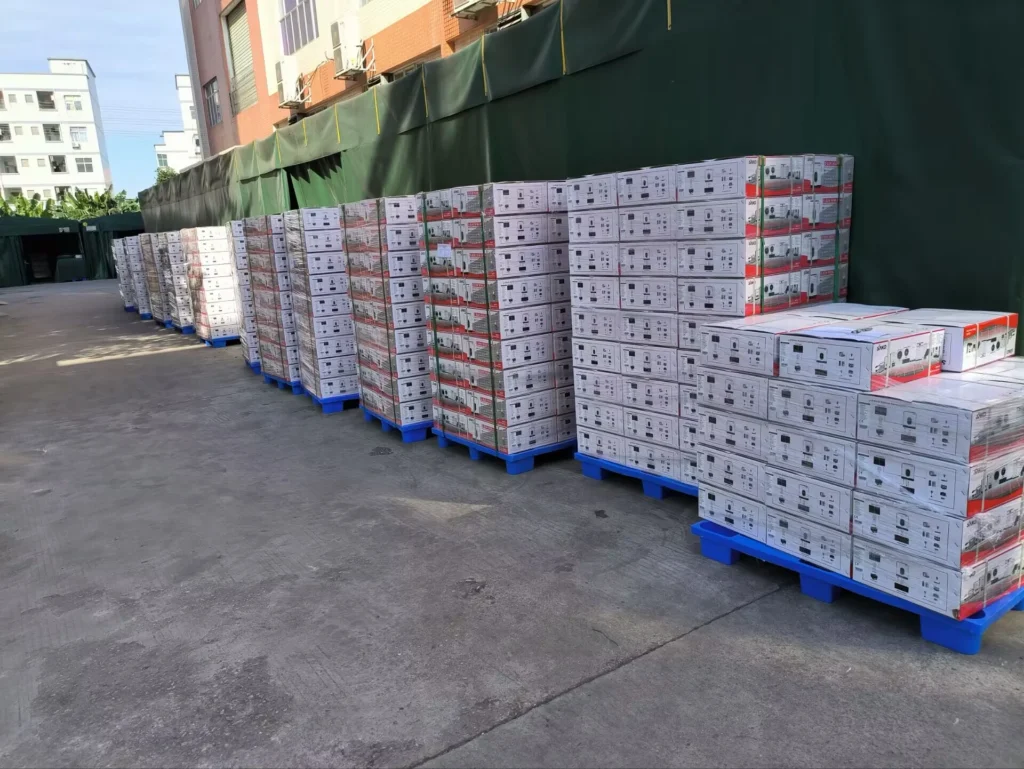Rows of stacked boxes on blue pallets outside a factory building with green tarps and industrial background.