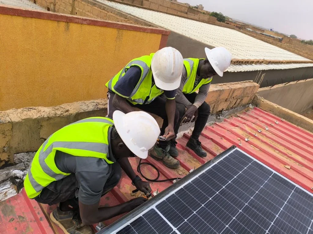 Three workers in high-visibility vests and helmets installing a solar panel on a red rooftop.